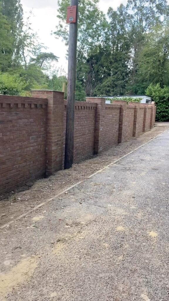 Finished red brick boundary wall with decorative dentil course and square piers alongside a gravel driveway.