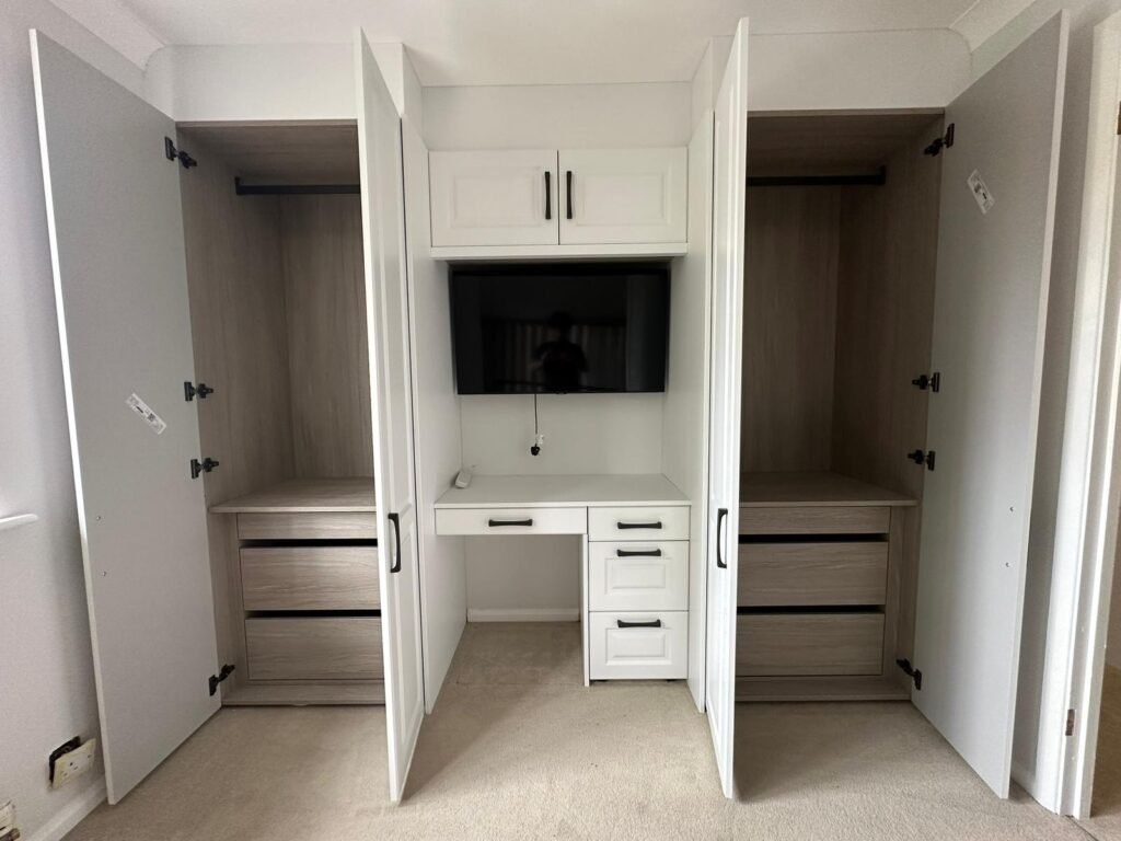 Custom white shaker bedroom storage unit with open oak-lined wardrobes, built-in desk, drawers, and wall-mounted television.