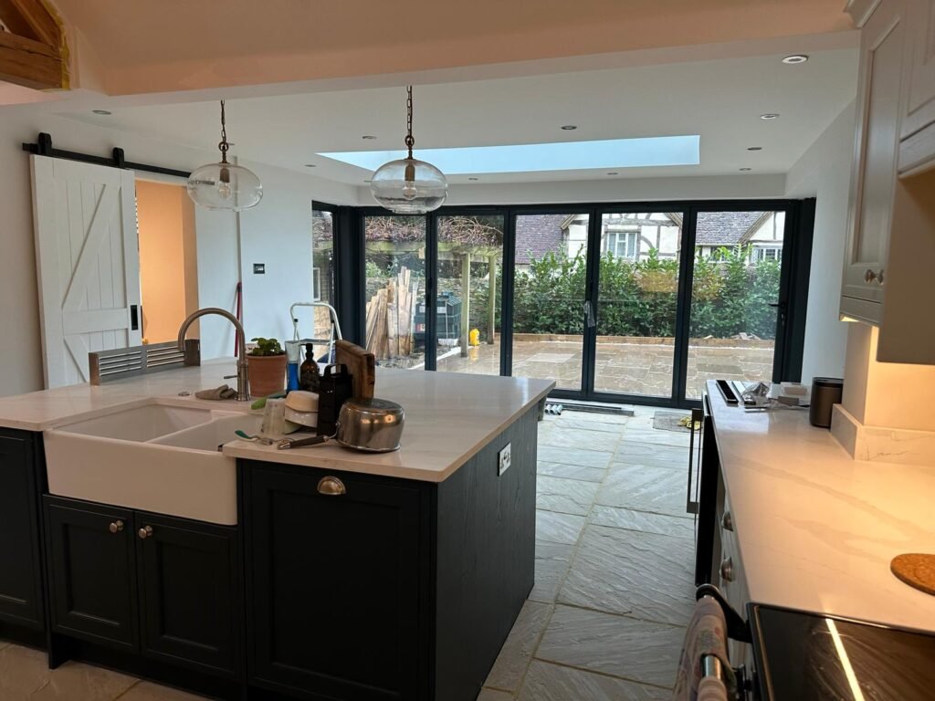 Finished open-plan kitchen with navy island, Belfast sink, bifold doors, roof lantern, and stone floor tiles.