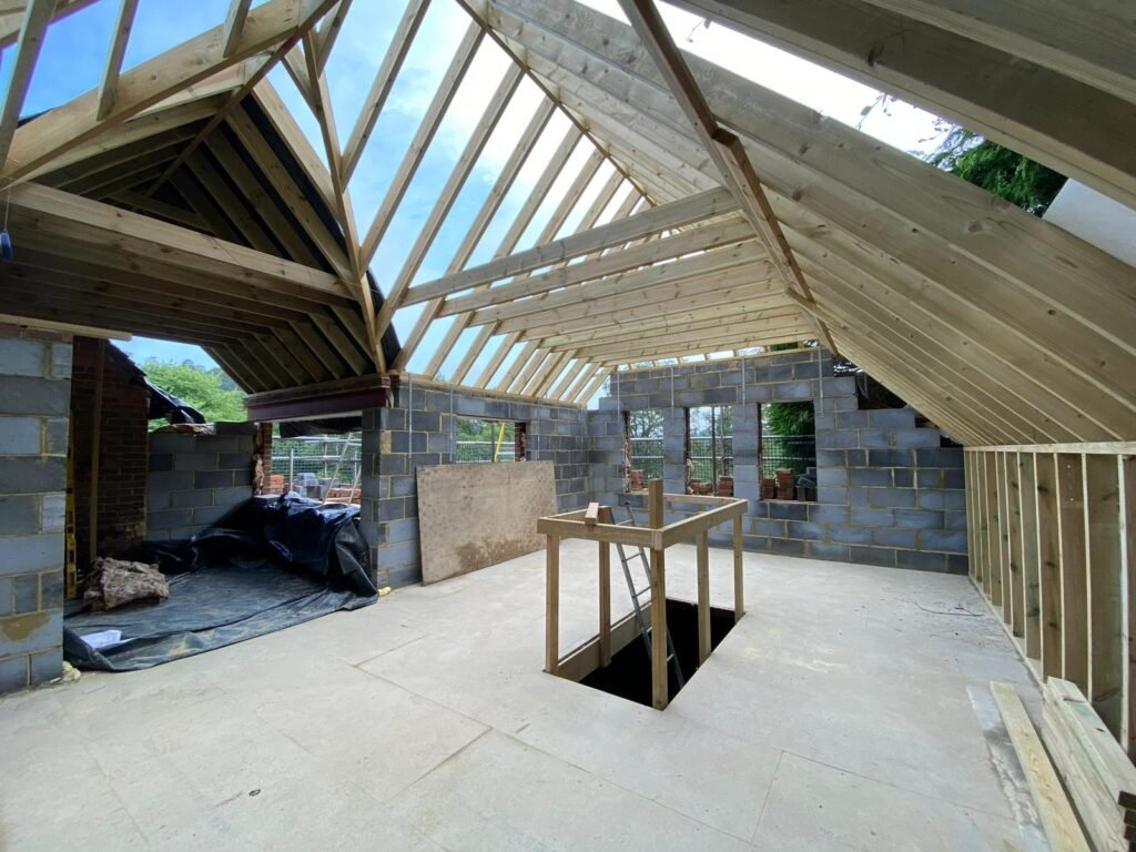 Interior loft conversion under construction showing exposed timber roof rafters, blockwork walls, and a staircase opening in the floor.