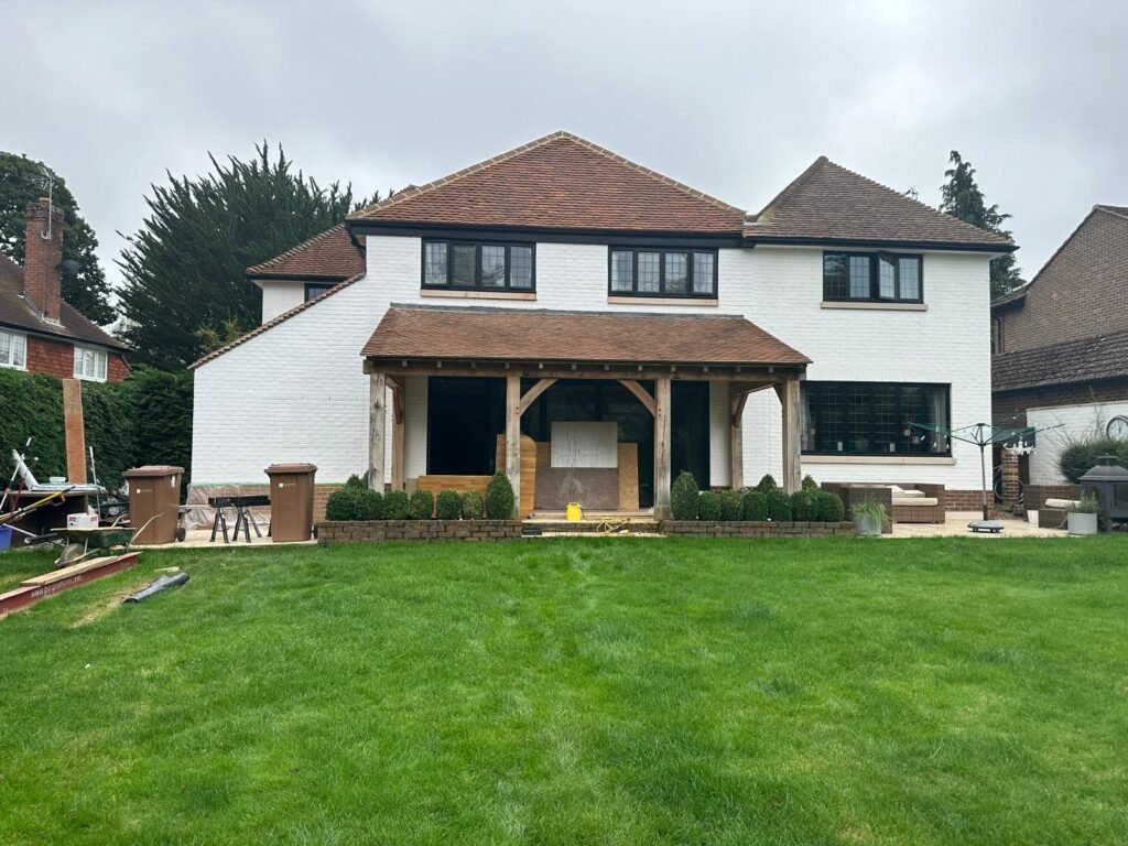 Wide rear view of a white brick house with a new oak timber porch, stone patio, and landscaped lawn.